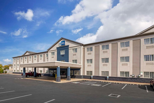 Three-story hotel exterior with a rectangular structure, large windows, and an entrance canopy. Multiple vehicles are parked in the lot under a partly cloudy sky.