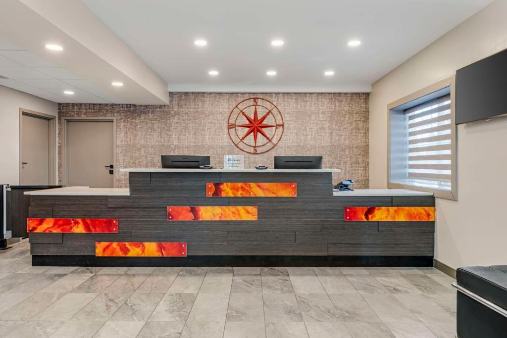 Modern hotel reception area with a dark wood front desk featuring illuminated orange panels, two computer screens, and a circular wall decoration. Marble tile floor and a window with blinds.