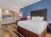 Hotel room with a large bed, purple sofa, side table, and a small kitchenette with a coffee maker. The walls are blue and white, and the floor has wooden panels.