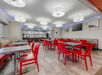 Bright dining area with red chairs, gray tables, and modern ceiling lights. Kitchen visible in the background.