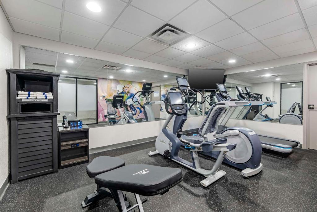 Small gym room with exercise machines, including ellipticals, a bench, and a mirror on the wall. Towels are stacked on a shelf in the corner.