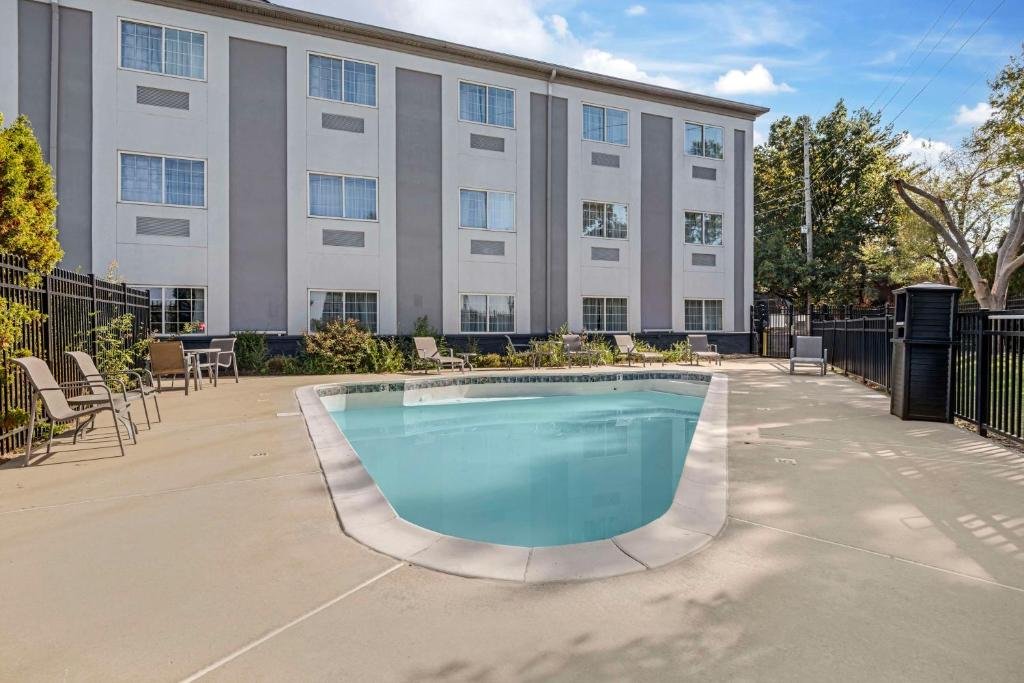 Outdoor pool area with surrounding chairs and tables, next to a multi-story building. Black fence encloses the space. Trees and shrubs are visible in the background under a blue sky.