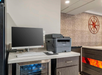 Office corner with a computer, printer, and mini fridge stocked with water bottles on a countertop. A decorative compass hangs on the wall nearby.