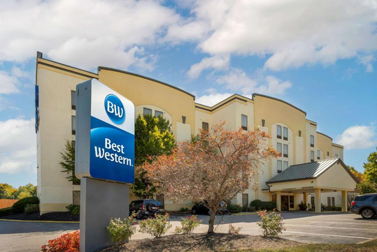 Exterior view of a Best Western hotel with a blue and white sign, showing a four-story building, some parked vehicles, and surrounding greenery under a partly cloudy sky.