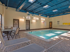 Indoor swimming pool area with a small pool, surrounded by lounge chairs and a round table. The ceiling is painted blue and walls have safety notices. Sunlight filters through windows.