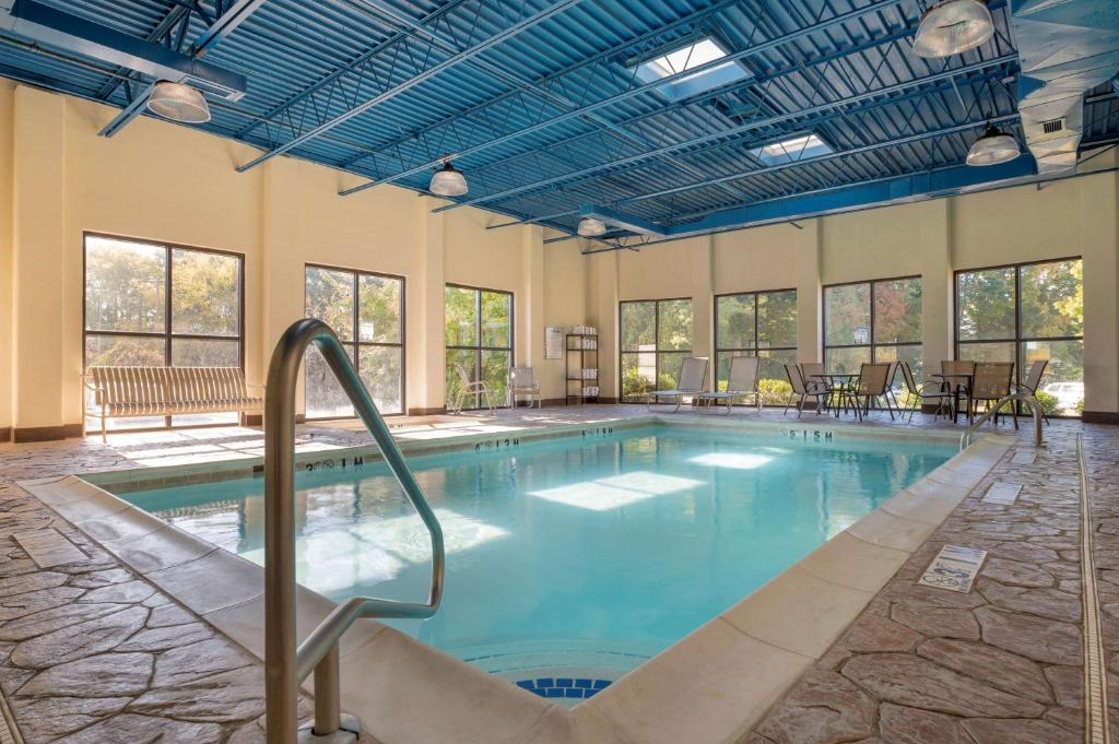 Indoor swimming pool with stone tile flooring, blue ceiling beams, large windows, and surrounding lounge chairs and tables. Natural light filters in through the windows.