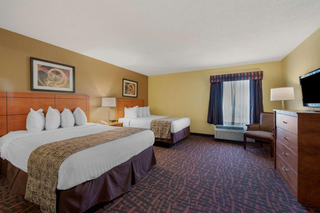 Hotel room with two double beds, a chair, a flat-screen TV on a dresser, and framed artwork on a beige wall. There's a large window with curtains, carpet flooring, and a couple of lamps.
