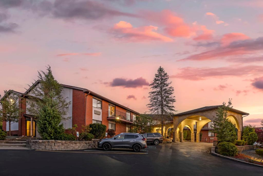 Two-story brick hotel building with an archway entrance at sunset. Several cars parked in front, surrounded by trees and shrubs under a colorful sky.