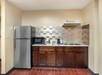 Compact kitchenette with wooden cabinets, stainless steel refrigerator, microwave, coffee maker, and a small stovetop. Tiled backsplash, hardwood floor.