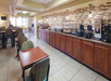 Hotel breakfast area with wooden tables and chairs. Buffet includes waffle makers, cereal dispensers, and a variety of food on a long counter. Tiled floor and decorative wall tiles.