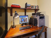 Home office setup with a wooden desk, desktop computer, lamp, and printer. Books are on a wall shelf above, with a chair in front of the desk.