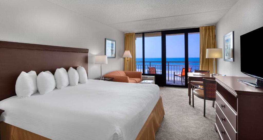 Hotel room with king-size bed, orange sofa, desk, and TV. Large windows with curtains open to reveal a balcony with ocean view. Neutral-toned decor and carpet.