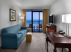 Hotel room with blue sofa, desk, and TV; balcony overlooking the ocean. Walls are white with a picture, and there's a floor lamp next to the sofa.