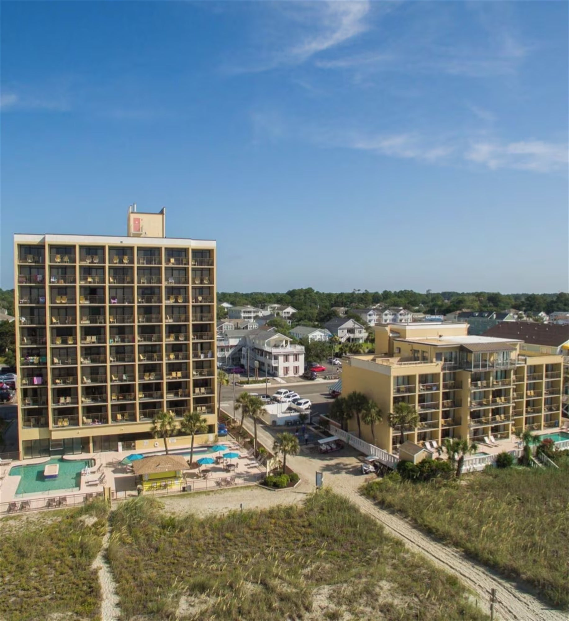 Two beige beachfront hotels with balconies and outdoor pools, surrounded by grass and small buildings, under a blue sky.