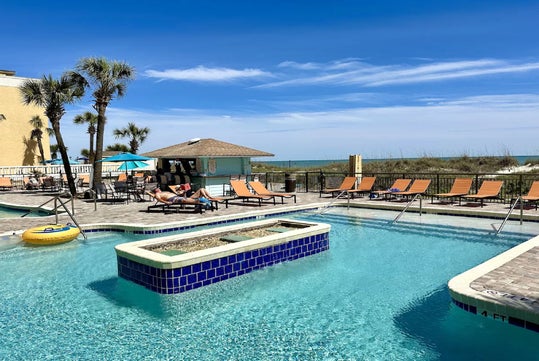 Outdoor pool area with lounge chairs and umbrellas, a person relaxing by the water, and a view of the beach and ocean in the background under a blue sky.
