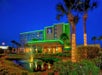 Hotel building with green neon lights, surrounded by palm trees and shrubs. A fountain is in the foreground under a deep blue evening sky.