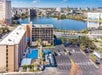 Aerial view of a hotel with a swimming pool, parking lot, and a lake in the background, set in an urban area under a clear blue sky.
