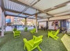 Covered outdoor area with green Adirondack chairs on artificial grass, near a pool. Tables and chairs are in the background, with some potted plants and a mural on the wall.