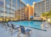 Outdoor hotel pool area with surrounding lounge chairs, framed by tall modern buildings and a clear blue sky.