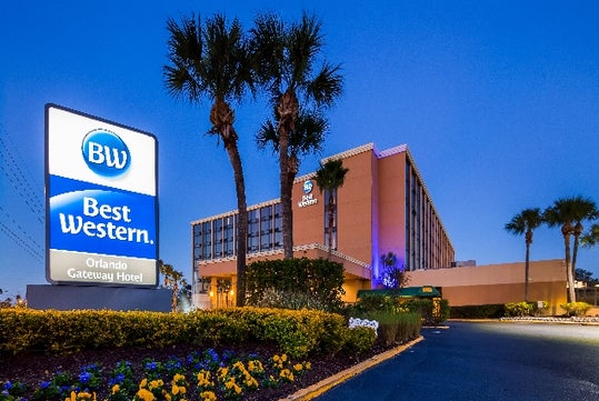 Best Western Orlando Gateway Hotel entrance at dusk, showing the hotel building, lit signage, and landscaped pathway with palm trees and flowers.