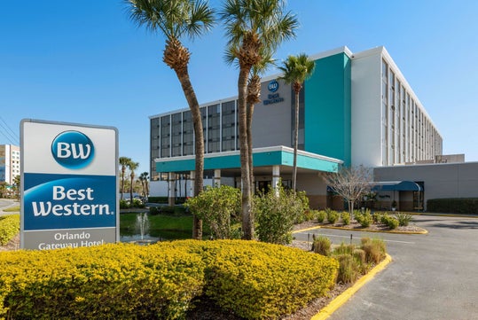 Exterior view of the Best Western Orlando Gateway Hotel with palm trees, landscaped bushes, and a visible hotel sign in front.