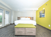 A bright bedroom with a large bed, green end bench, and hardwood floors. Two doors with windows on the left; colorful waterfall painting on a yellow accent wall to the right.