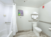 A modern bathroom with a bathtub, shower curtain, toilet, and towel rack. The walls are white with a framed picture, and there are safety bars near the toilet and bathtub.
