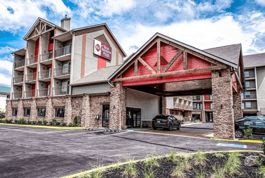 Exterior view of a multi-story hotel with stone accents and a covered entrance, featuring a logo on the building. A parked car is near the entrance, with a cloudy sky above.
