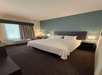 A hotel room with a large bed, two bedside tables with lamps, an armchair, and a window with curtains. The walls are painted white and teal, and the floor is carpeted.