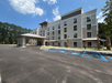 A four-story hotel building with a modern facade, surrounded by trees. The parking lot includes marked accessible spaces. The sky is partly cloudy.