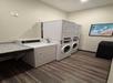 Laundry room with a stackable washer and dryer, a separate dryer, a countertop, and an ocean-themed picture on the wall. The floor is wooden, and there is a black trash bin.