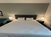 A neatly made bed with a dark headboard, flanked by two bedside tables with lamps. A teal armchair is partially visible on the left.