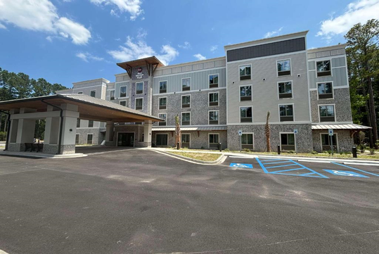 A four-story hotel with a stone and siding exterior, featuring numerous windows and a covered entrance. The foreground shows a parking lot with marked spaces under a partly cloudy sky.