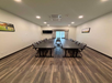 A rectangular conference room with a long table and twelve chairs. There is a wall-mounted TV and framed pictures on the walls. The floor is wood-paneled.