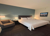Hotel room with a large bed, two bedside lamps, a gray armchair, and a framed picture on the wall. The room has a carpeted floor and a blue accent wall behind the bed.