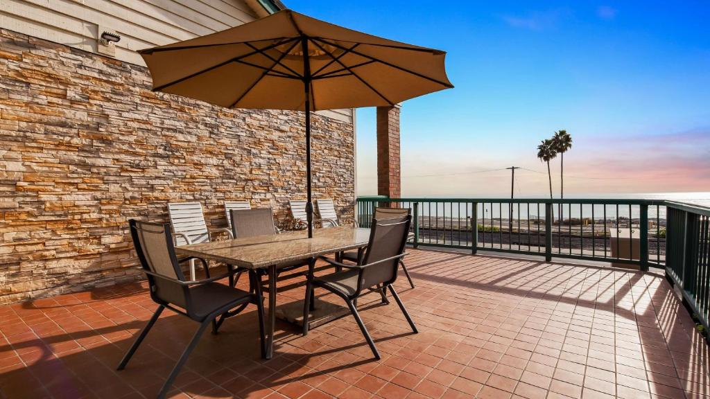 Patio with a stone wall, table, chairs, and large umbrella on tiled flooring. Overlooks ocean with two tall palm trees visible against a clear blue sky.