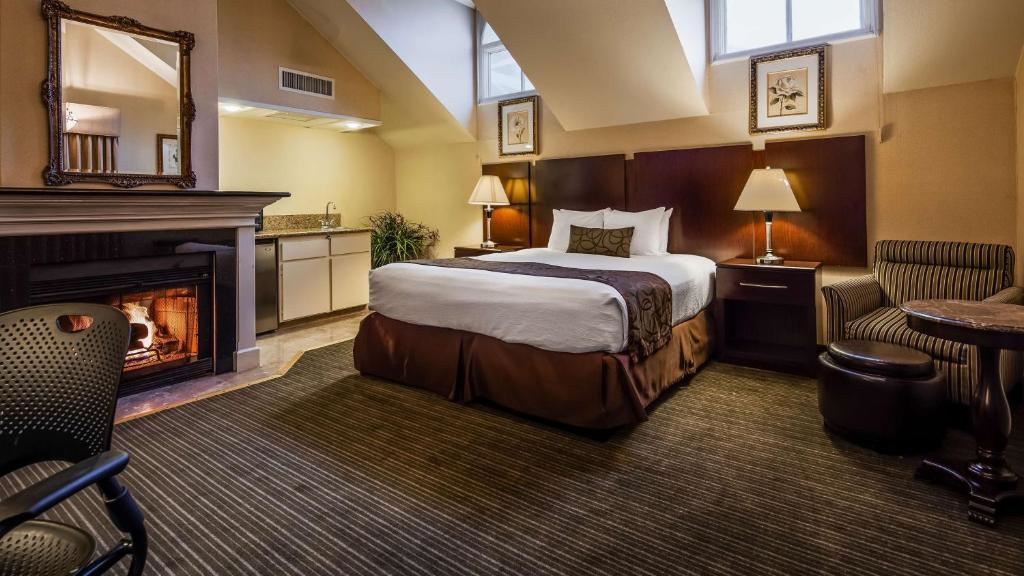 A cozy hotel room features a bed, armchair, small table, fireplace, and kitchenette. Two skylights and framed pictures adorn the cream-colored walls, creating a warm atmosphere.