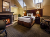 A cozy hotel room features a bed, armchair, small table, fireplace, and kitchenette. Two skylights and framed pictures adorn the cream-colored walls, creating a warm atmosphere.