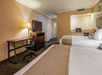 Hotel room with two double beds, a desk with a chair, a TV on a dresser, and a small kitchenette in the corner. Walls are tan-colored and decor is minimal.