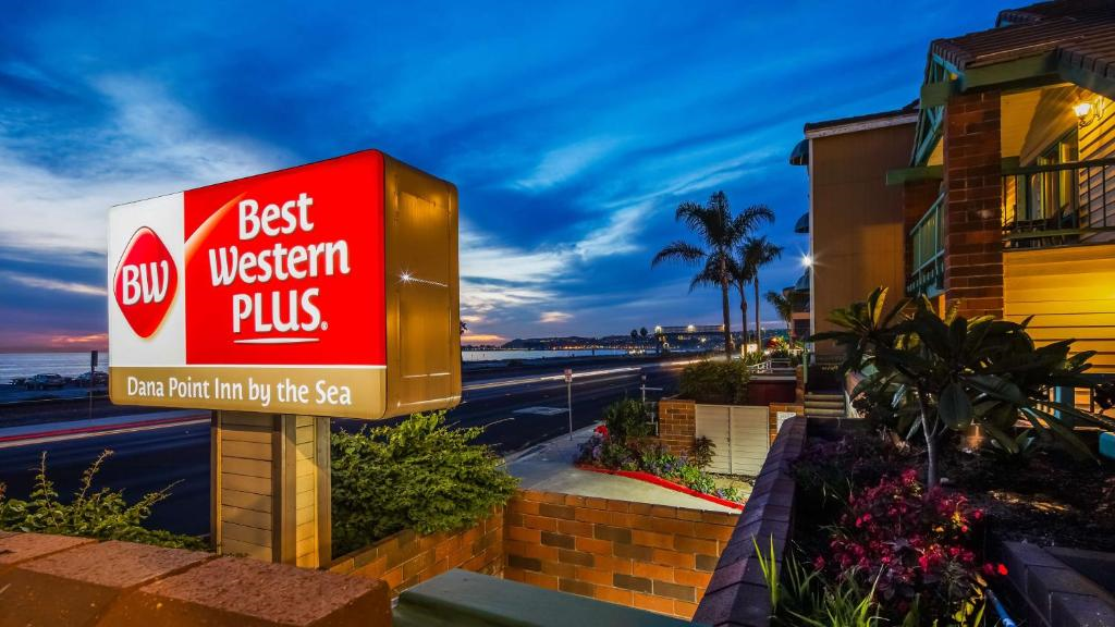 Best Western Plus Dana Point Inn by the Sea sign illuminated at dusk with a view of the street, palm trees, and ocean in the background.