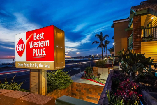 Best Western Plus Dana Point Inn by the Sea sign illuminated at dusk with a view of the street, palm trees, and ocean in the background.