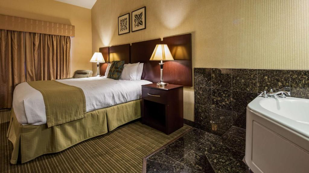 Hotel room with a large bed, two bedside lamps, framed art, and a jacuzzi in the corner. The decor features brown and beige tones. A window with curtains is on the left.