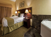 Hotel room with a large bed, two bedside lamps, framed art, and a jacuzzi in the corner. The decor features brown and beige tones. A window with curtains is on the left.