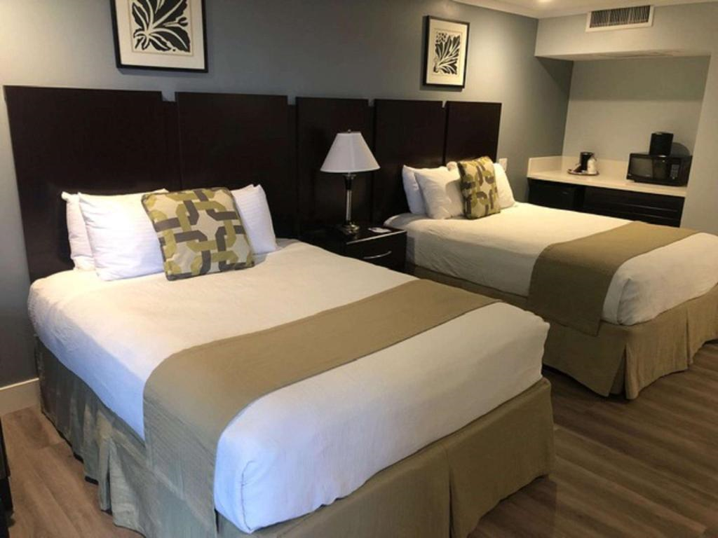 Hotel room with two double beds, each with white sheets and beige throws. A lamp and wall art are visible between the beds, and a small kitchenette with appliances is in the background.