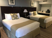 Hotel room with two double beds, each with white sheets and beige throws. A lamp and wall art are visible between the beds, and a small kitchenette with appliances is in the background.