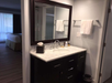Hotel room bathroom with a dark wood vanity, marble countertop, large mirror, tissues, and towels. Bedroom visible in the background through an open doorway.