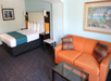 Hotel room with a king bed, teal accent runner, orange sofa, glass coffee table, and framed wall art on light blue walls.