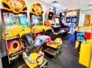 Room filled with various arcade games, including racing simulators, classic arcade cabinets, and a partially visible air hockey table on the right.