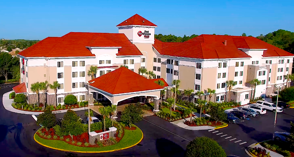 A large hotel building with white walls and red roofs is surrounded by palm trees, landscaped gardens, and a parking lot with several vehicles.