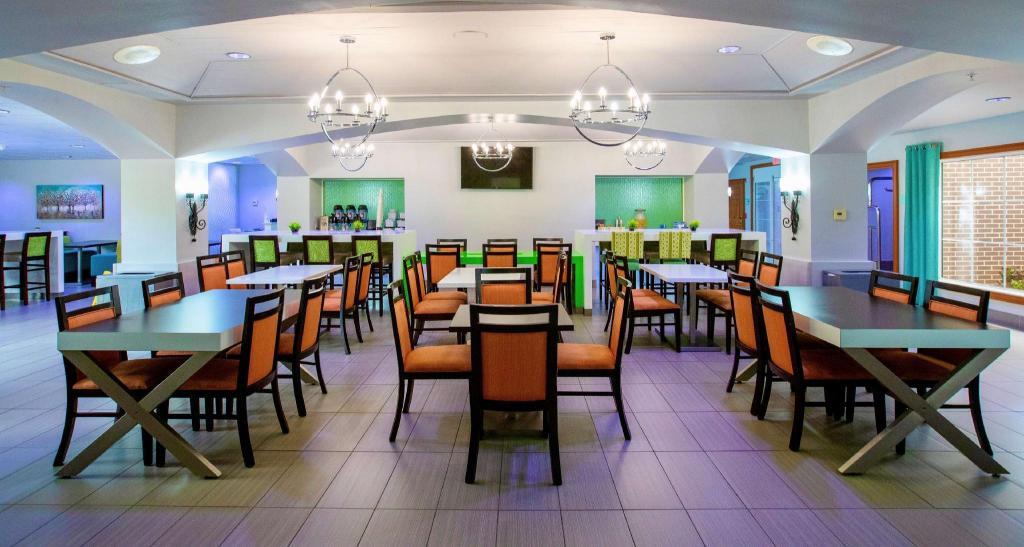 A modern dining area with square tables and orange-cushioned chairs, arranged in rows. The room has tiled floors, chandeliers, and a buffet counter at the back.